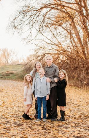 A family group poses together outdoors on fallen autumn leaves against a backdrop of bare trees at sunset.