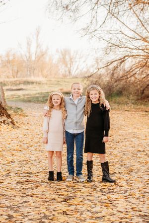 Three children stand together on a path covered in fallen autumn leaves during a golden sunset.