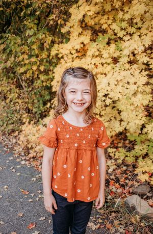 A smiling child in an orange polka dot top stands against golden autumn foliage on a sunny afternoon.