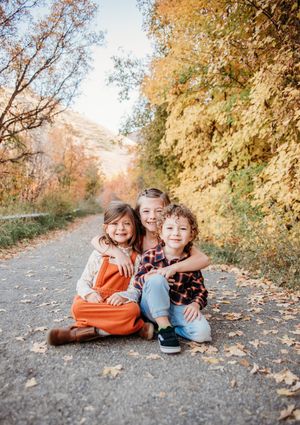 Young children sit together on a leaf-covered path surrounded by vibrant autumn foliage on a sunny fall day.
