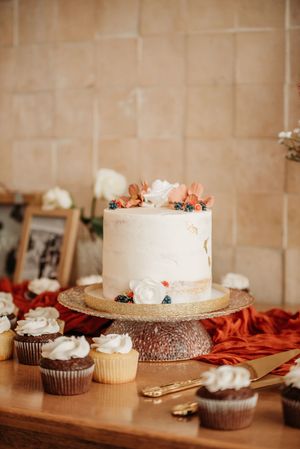 White wedding cake and chocolate cupcakes displayed on rustic table with orange decor accents.