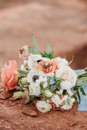 A delicate bridal bouquet with coral and white roses, anemones and greenery rests on red desert rock.