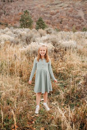 Young girl in a blue-gray dress stands smiling in a field of tall dried grass during autumn.