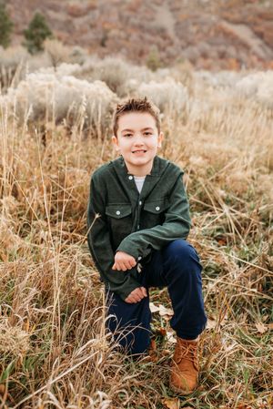 A child wearing a green jacket and brown boots sits in a field of tall autumn grass.