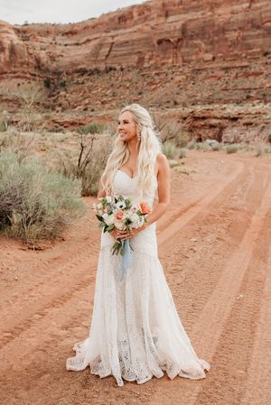 Delicate lace wedding dress captured against stunning red rock canyon landscape on dusty desert trail.