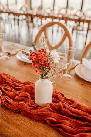 Close-up of rustic table decoration featuring white vase with coral flowers and draped terracotta fabric runner.