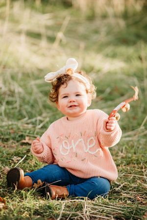 A small child in a peach sweater and jeans sits on grass during golden hour.