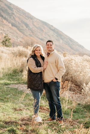 Two people share a sweet moment together on a grassy hillside with mountains in the background at sunset.