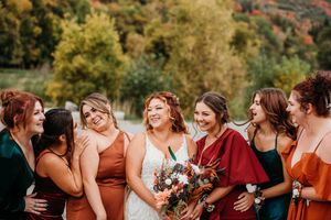Bridal party in warm autumn colors of burgundy and rust orange share candid laughter at an outdoor wedding celebration.