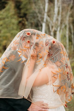 A white sheer veil with delicate peach floral embroidery patterns billowing in the wind.