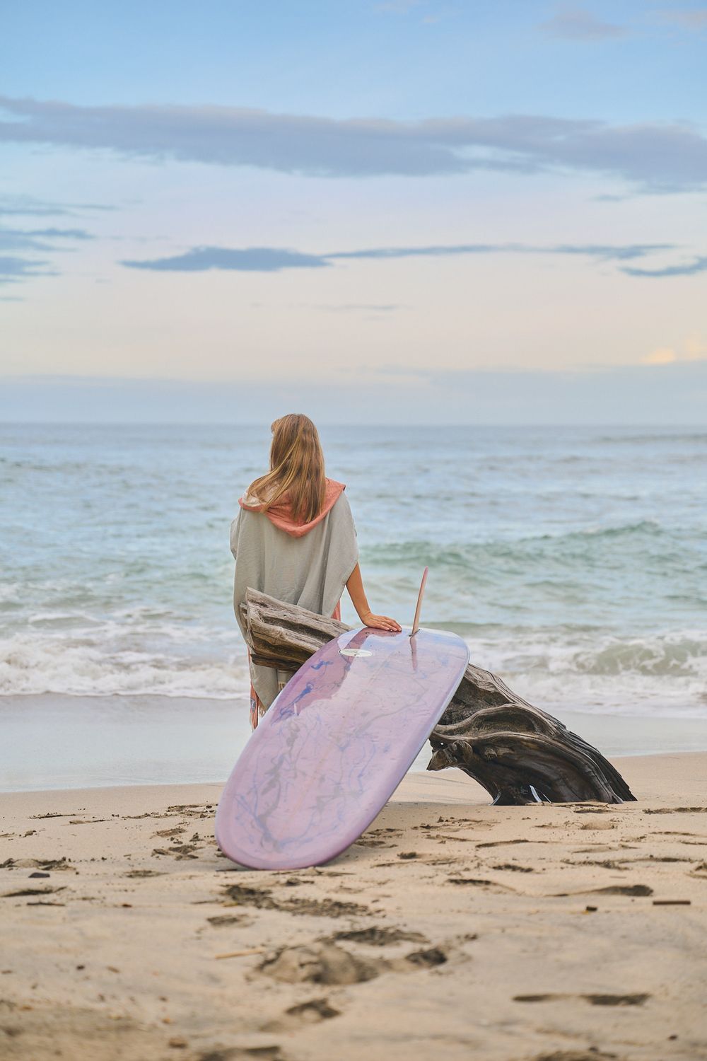 A surfer holding a purple surfboard crouches on the sandy beach at sunset with waves and clouds in the background.