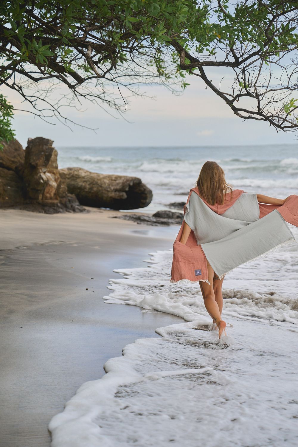 A woman in a peach-colored dress walks along a sandy beach with gentle waves and fallen tree branches.