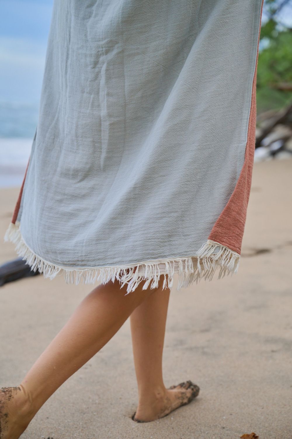 A person wearing sandals walks on sand with the edge of a white linen dress visible in a motion sequence.