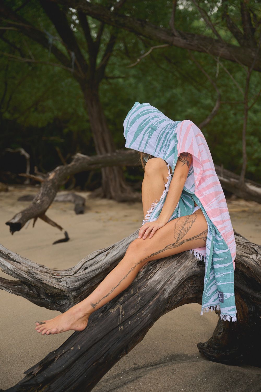 Bare feet resting on a weathered driftwood log on a sandy beach, with a colorful striped beach towel draped nearby.