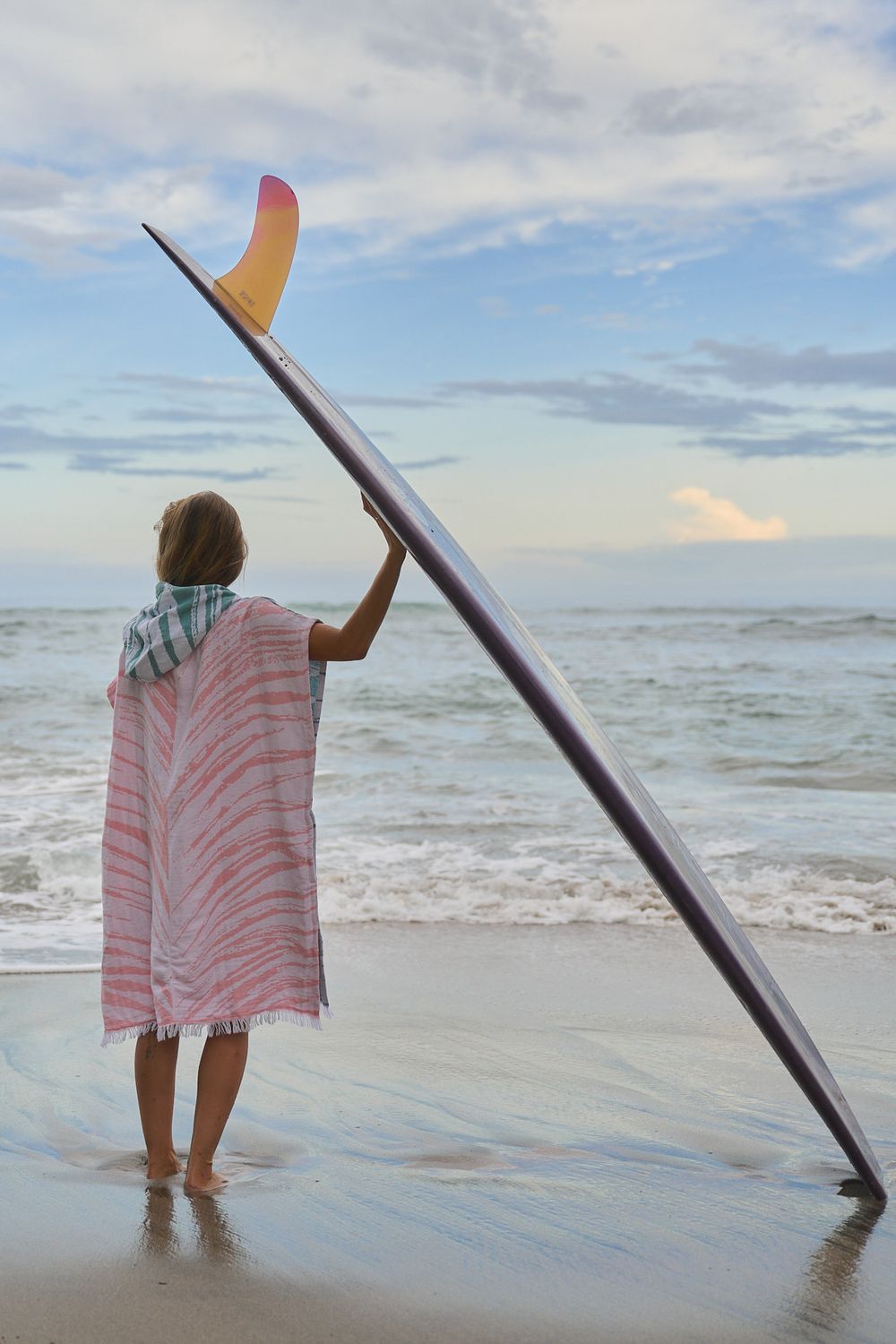 A surfer wrapped in a pink towel stands at the ocean's edge with a surfboard at sunset.