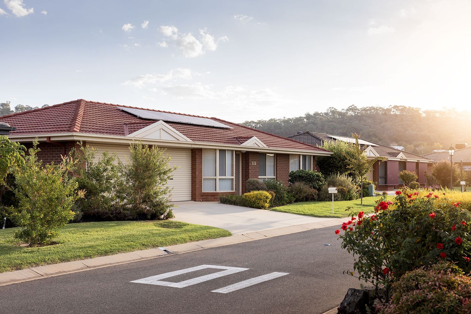 Modern suburban house with red tiled roof and manicured lawn on residential street at sunset.