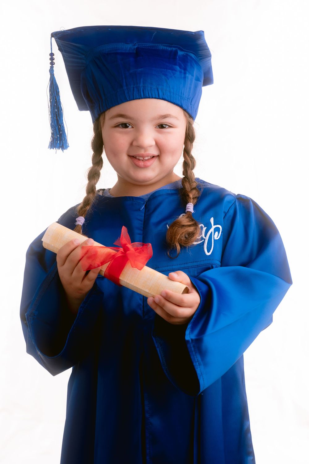 Smiling graduate wearing blue cap and gown displaying diploma with red bow against white backdrop.