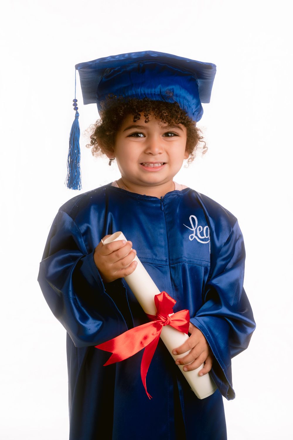 Young graduate wearing a blue cap and gown holds a diploma with red ribbon during graduation ceremony against white backdrop.