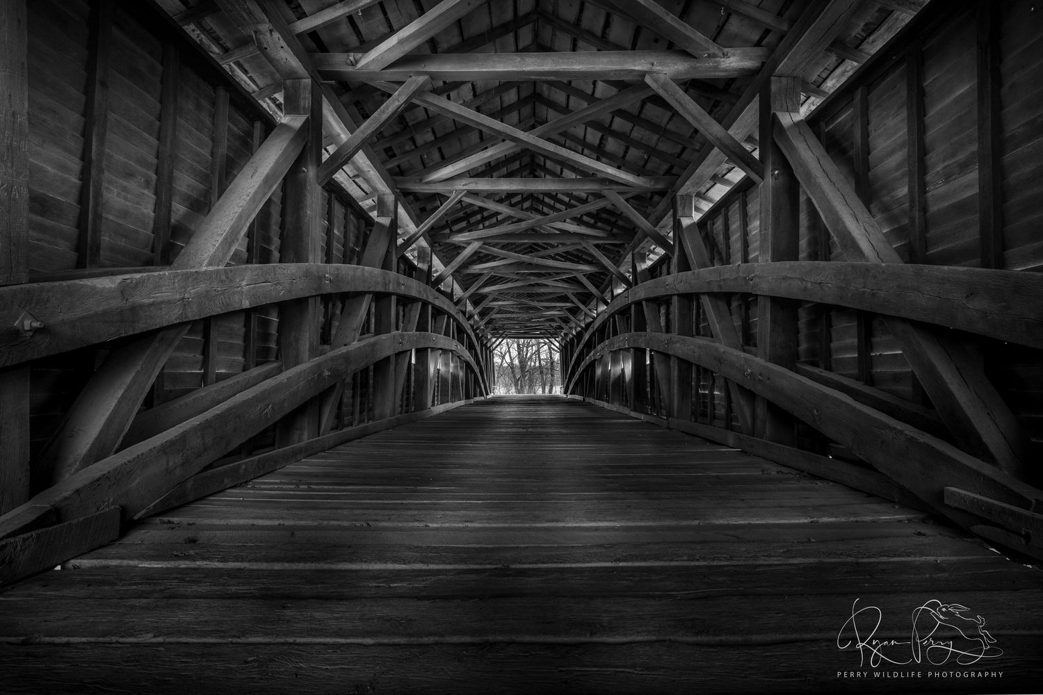 Ancient covered wooden bridge spans river with exposed support beams and wooden planks in black and white.