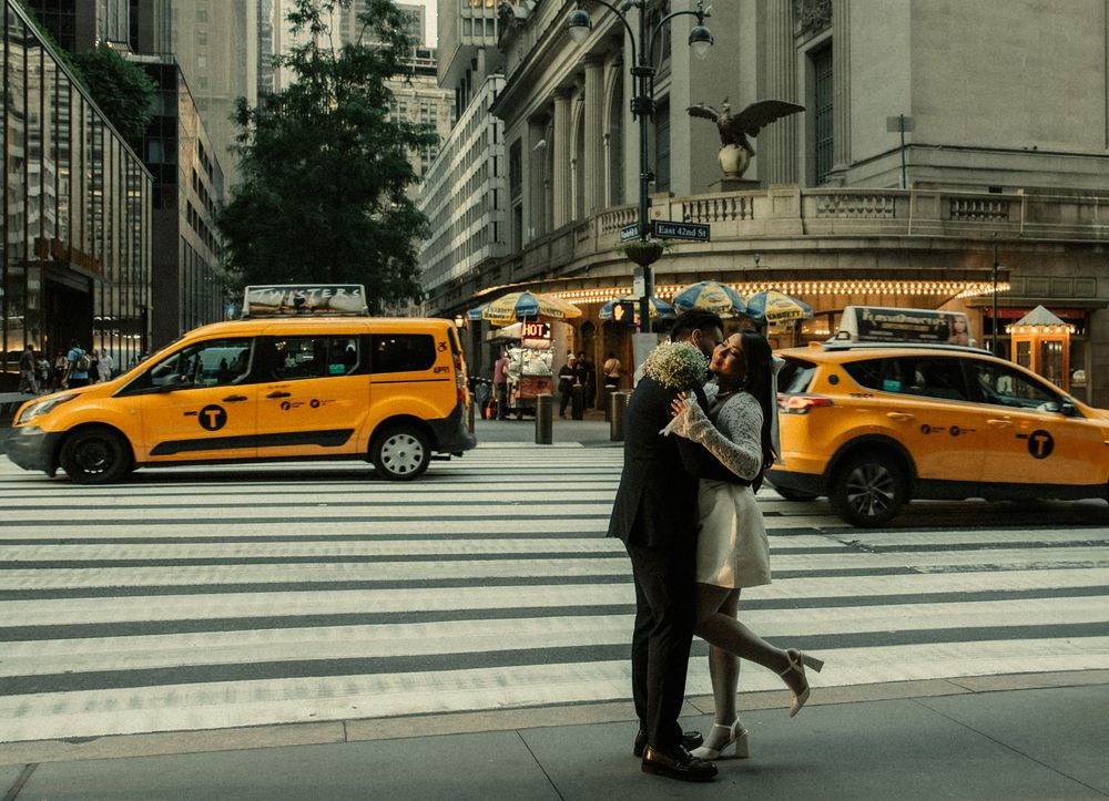 Yellow taxi cabs line a busy street corner in New York City while people cross at a crosswalk at dusk.