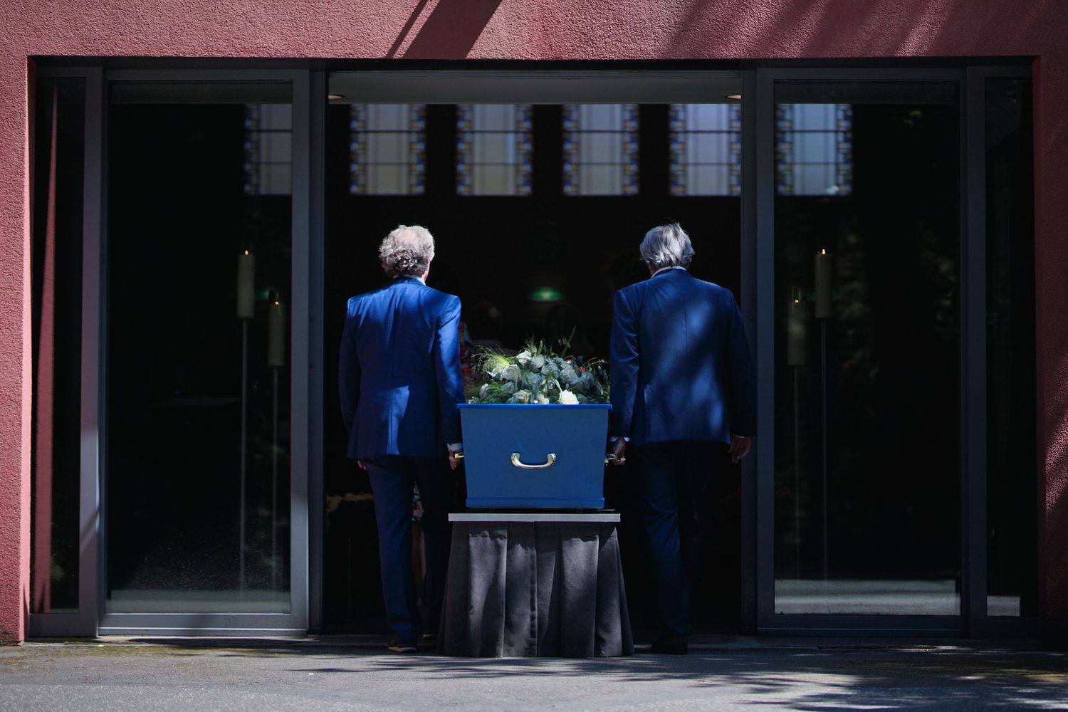 Funeral pallbearers in blue suits carrying casket through doorway.