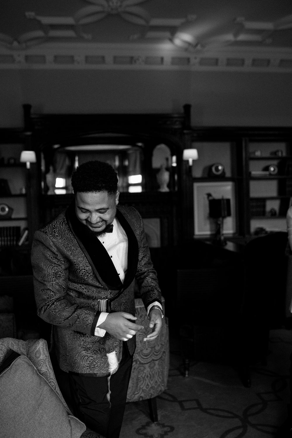 Groom in ornate patterned tuxedo jacket smiling while adjusting attire in an elegant dark interior room.