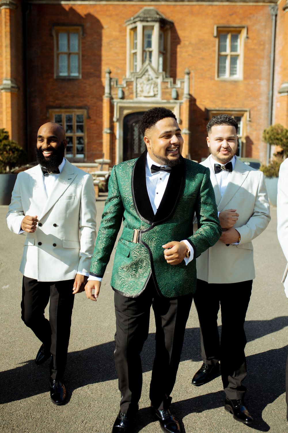 Groom in green brocade jacket with groomsmen in white tuxedos outside a grand brick wedding venue.