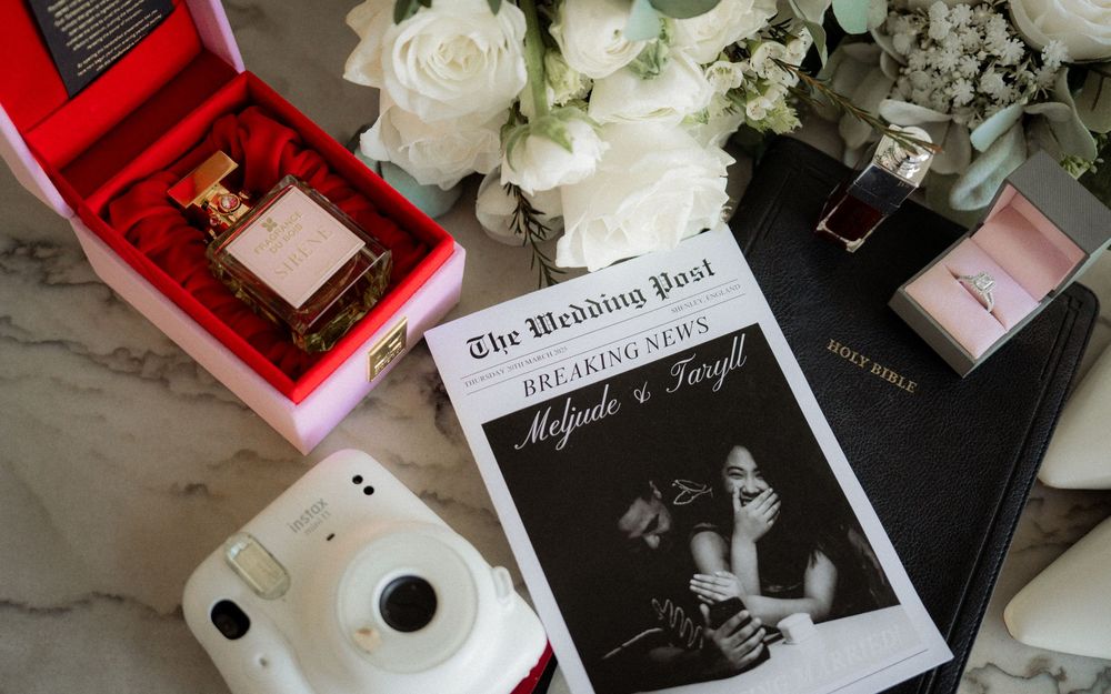 Flat lay of wedding details including perfume, ring, Bible, Instax camera, and custom wedding newspaper.