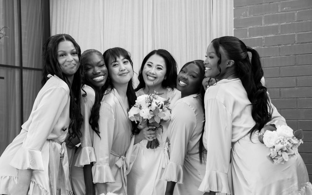 Black and white photo of bride with five bridesmaids in robes smiling and holding a floral bouquet.