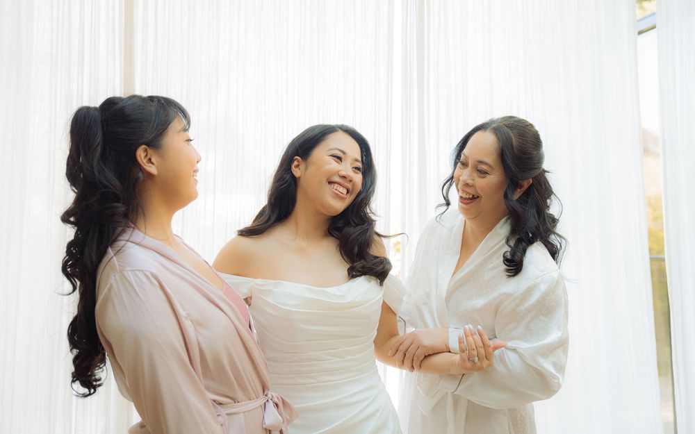Bride in white gown laughs with two bridesmaids in robes during wedding morning preparations.