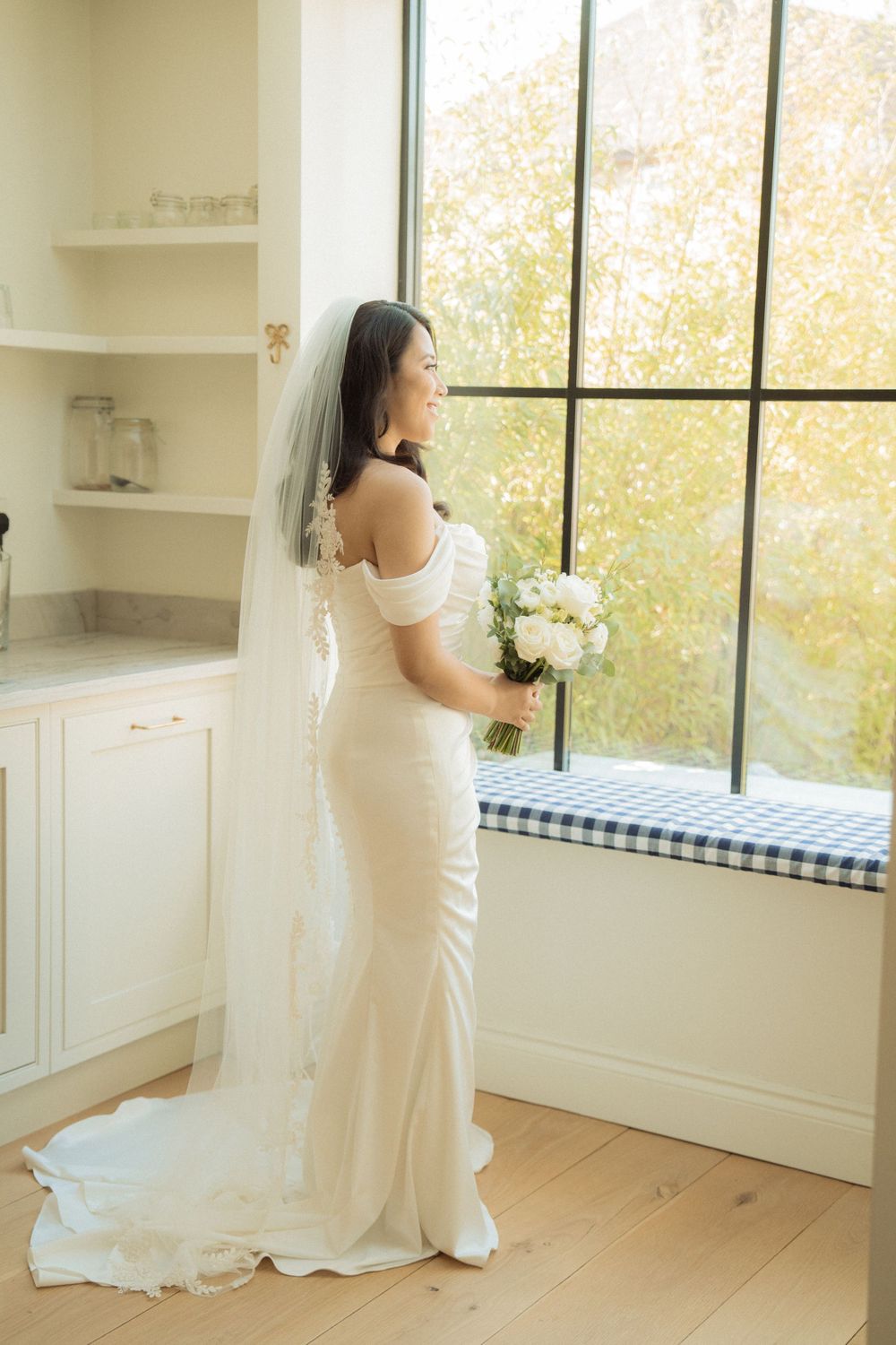 Bride in fitted white gown and veil gazes out large window holding white floral bouquet.