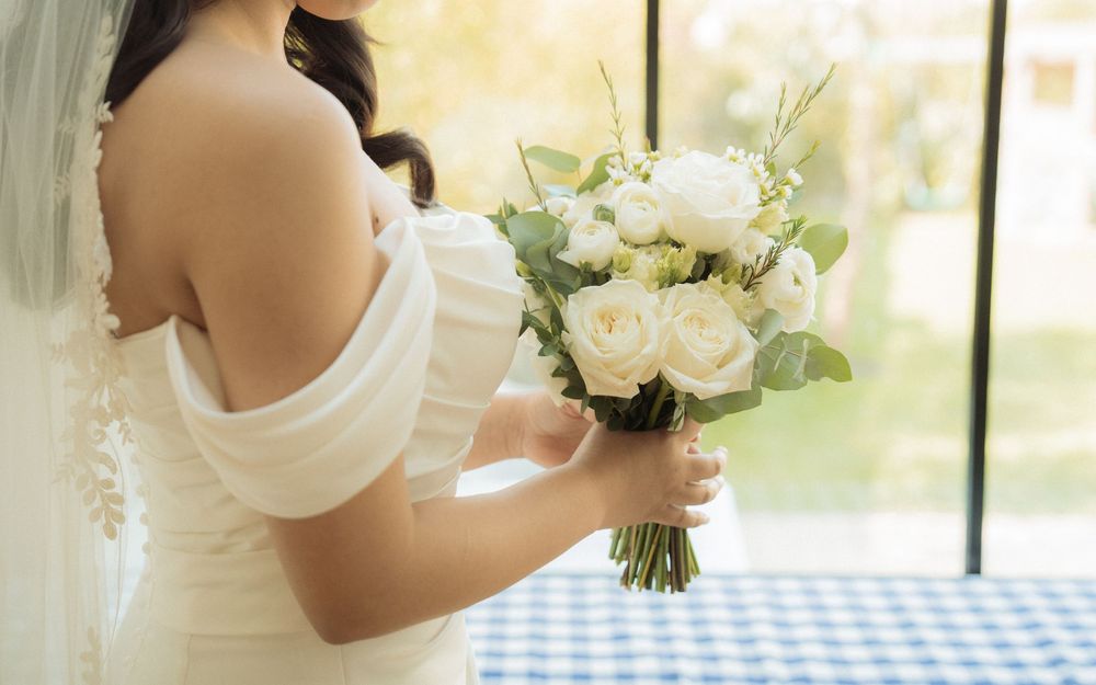 Bride in off-shoulder white gown holds lush white rose and eucalyptus wedding bouquet.