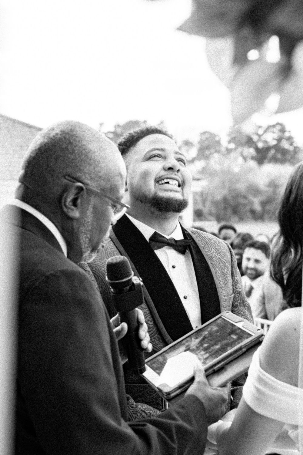 Groom laughing joyfully during wedding ceremony while officiant holds microphone in black and white photo.