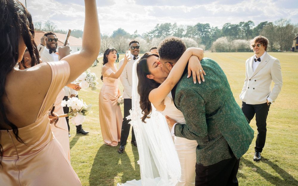 Bride and groom share first kiss surrounded by cheering wedding party on sunny outdoor lawn.