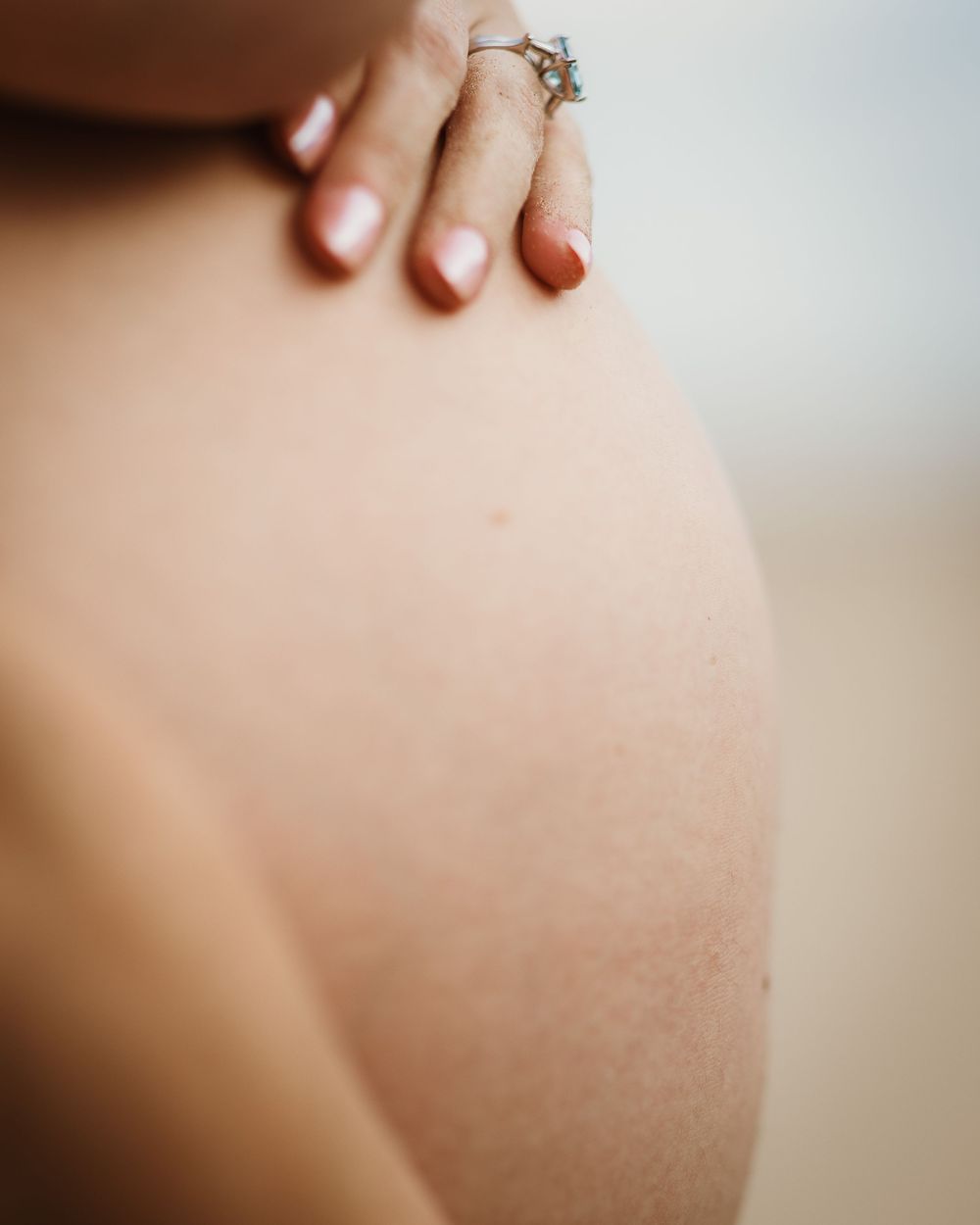 Close-up of a person's bare shoulder with a hand resting on it, wearing a delicate ring with red nails.