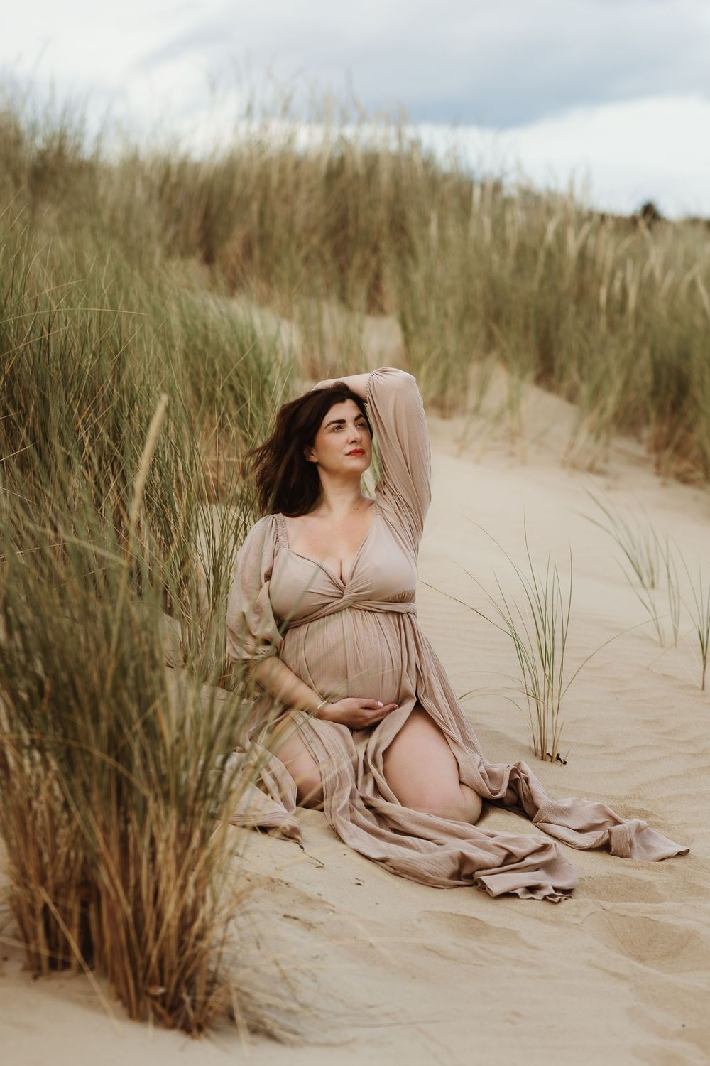 Pregnant woman in beige dress sitting on sandy dunes surrounded by tall grass during an outdoor maternity photoshoot.