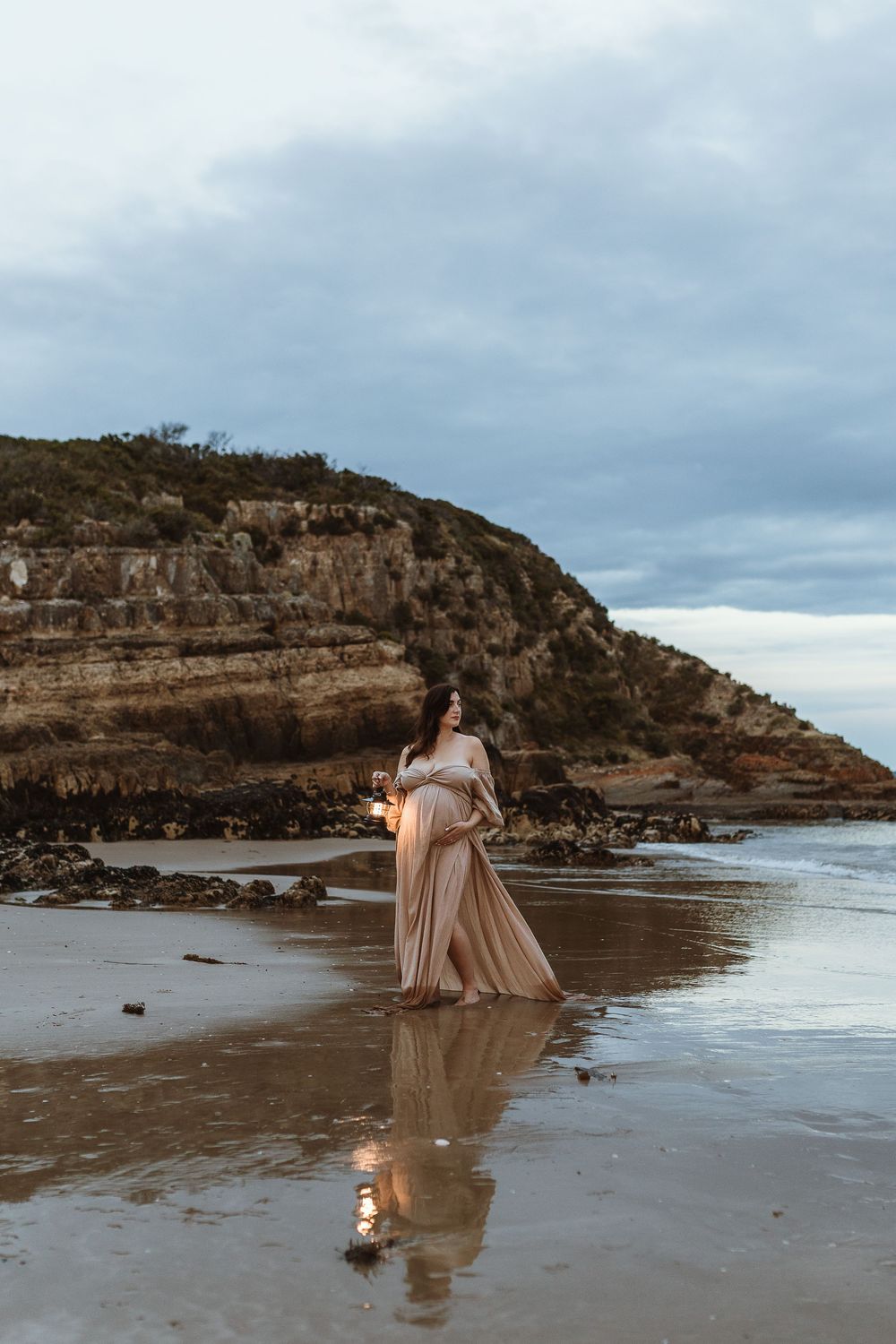 Pregnant woman in flowing beige gown standing on a reflective beach with rocky cliffs under a cloudy sky.