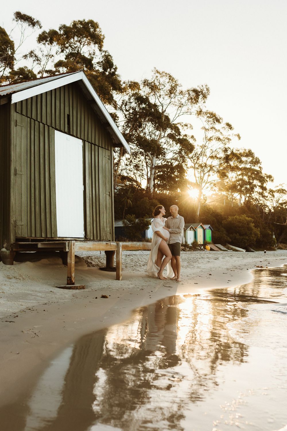 Couple embracing at sunset on sandy beach beside rustic wooden boat shed with colorful beach boxes.
