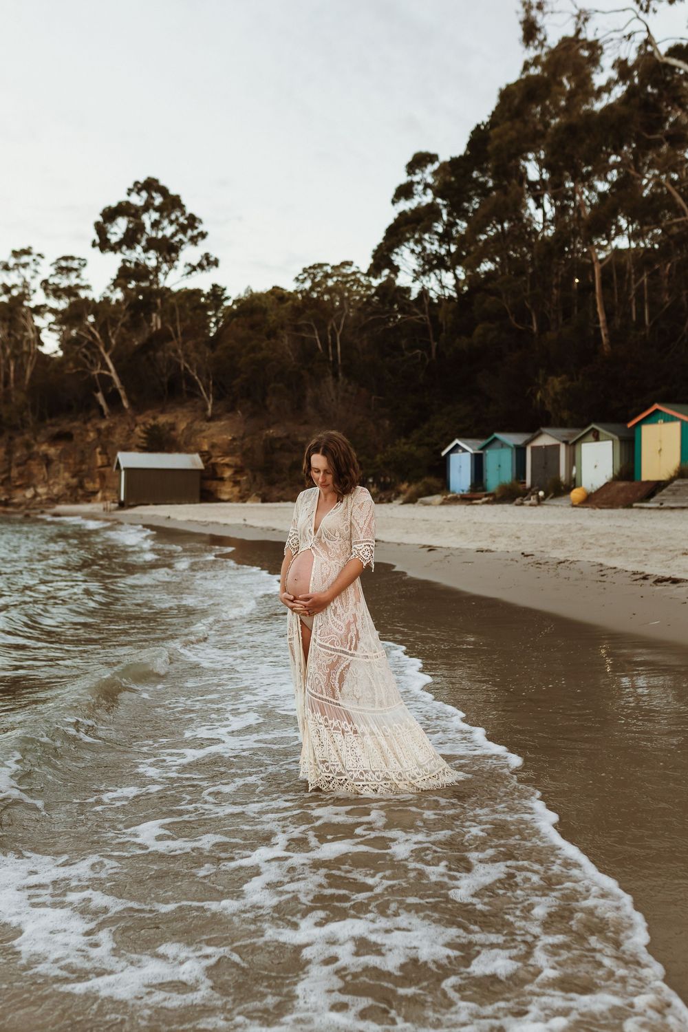 Pregnant woman in white lace dress standing in ocean waves on sandy beach with colorful beach huts in background.