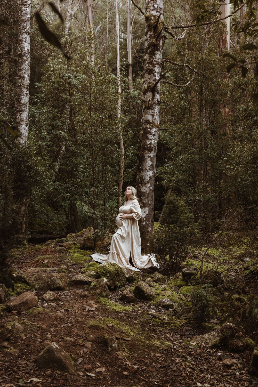 Pregnant woman in white gown standing among mossy rocks in a misty forest, surrounded by tall trees.