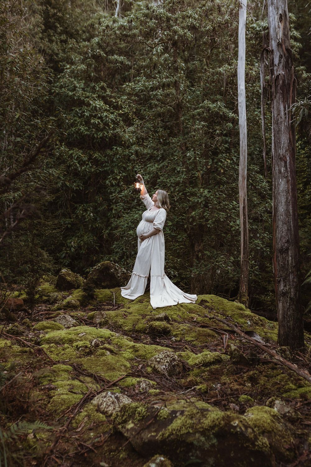 Pregnant woman in white gown standing on mossy rocks in lush forest, reaching up toward light filtering through trees.