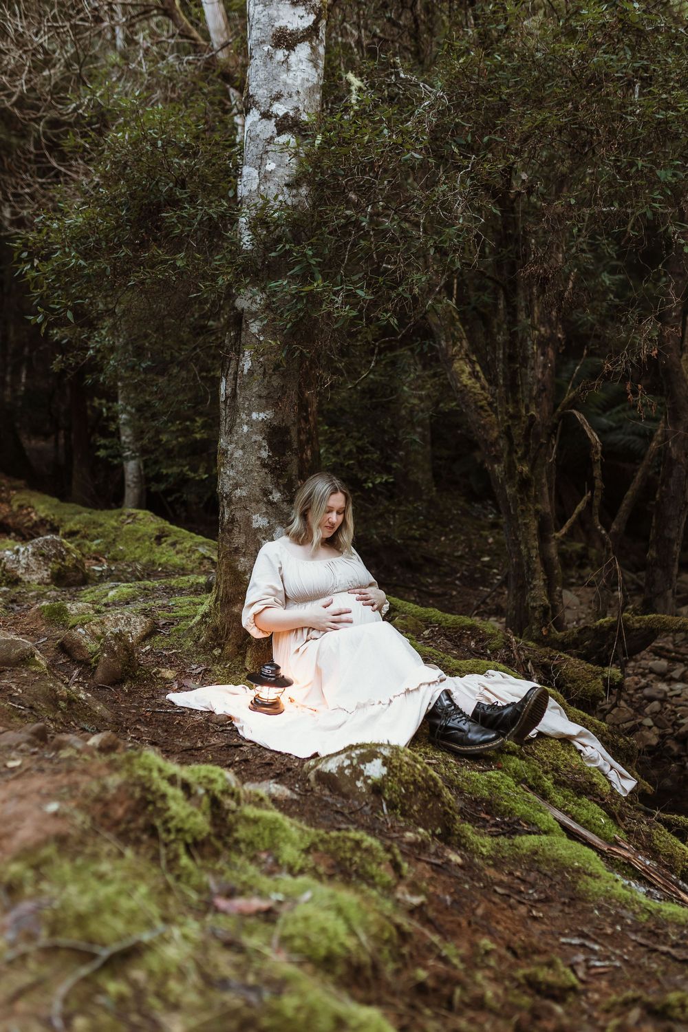Pregnant woman in white dress sitting on mossy rocks in a lush green forest during a maternity photoshoot.