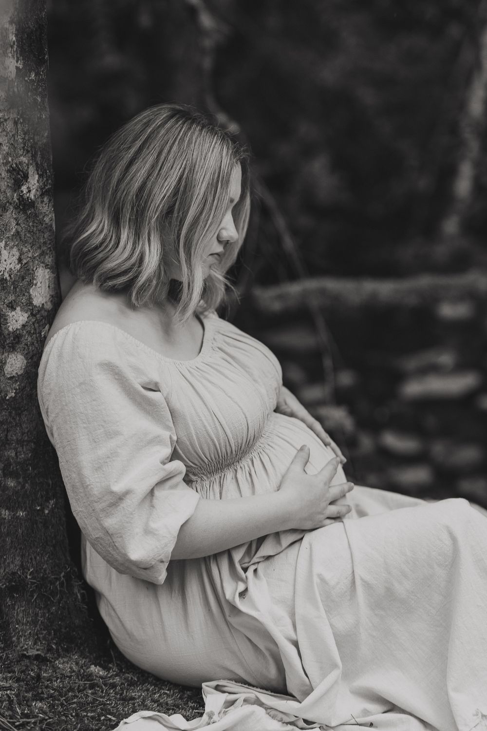 Black and white maternity photo of a pregnant woman in a flowing dress sitting against a tree, cradling her baby bump.