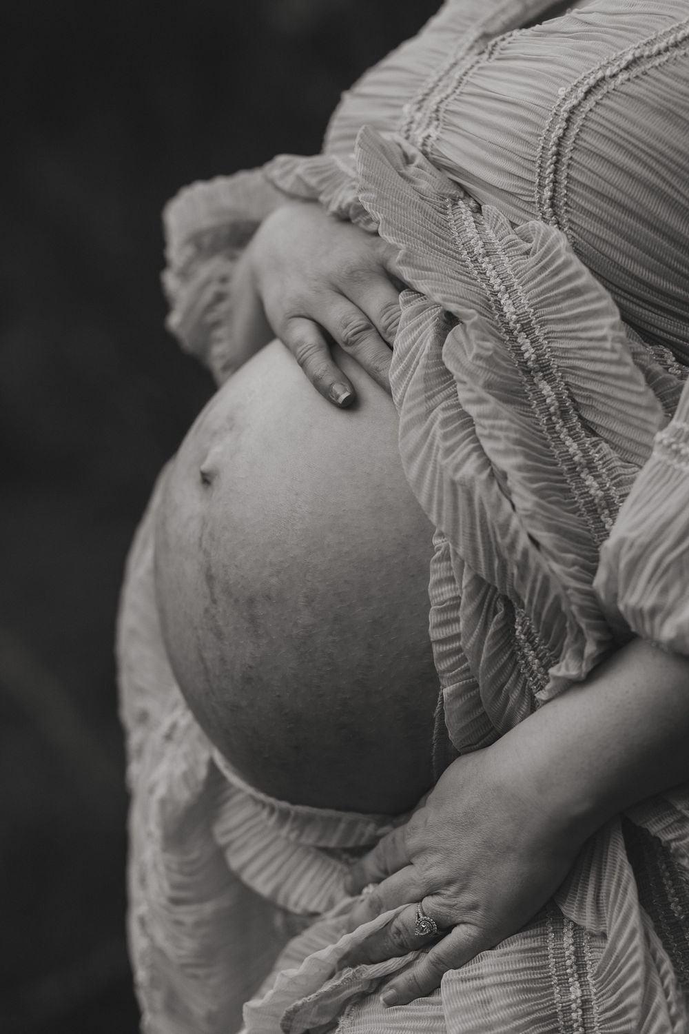 Black and white maternity photo of a pregnant woman cradling her bare baby bump in a ruffled dress.