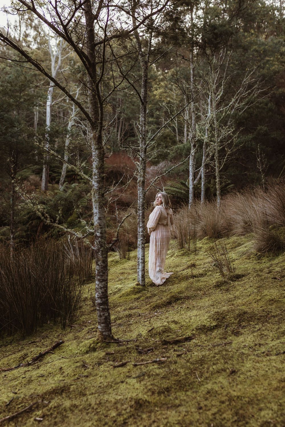 Pregnant woman in flowing blush dress standing among birch trees on mossy hillside in forest.