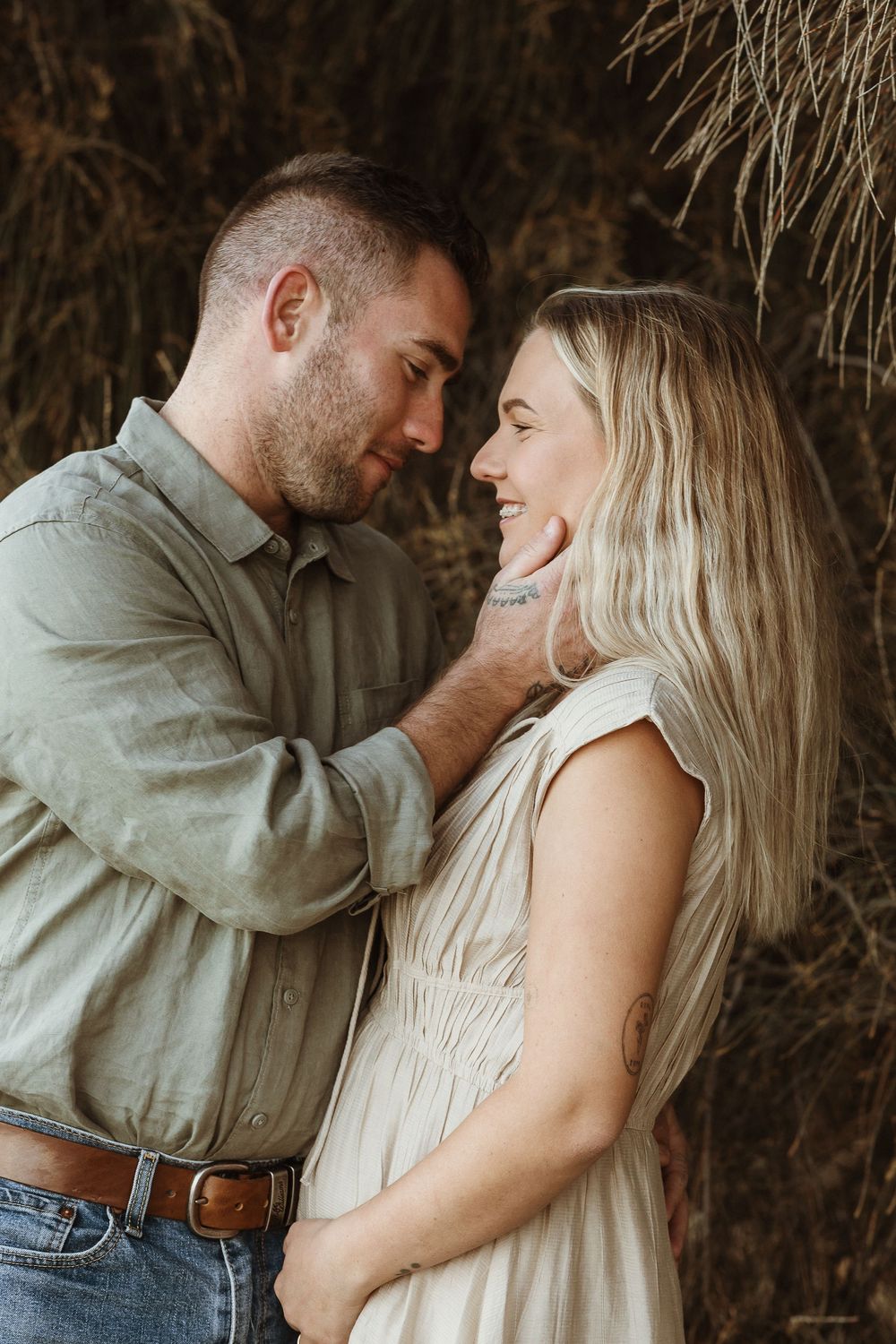 A couple shares a tender moment, the man gently holding the woman's face as they gaze at each other near dried grass.