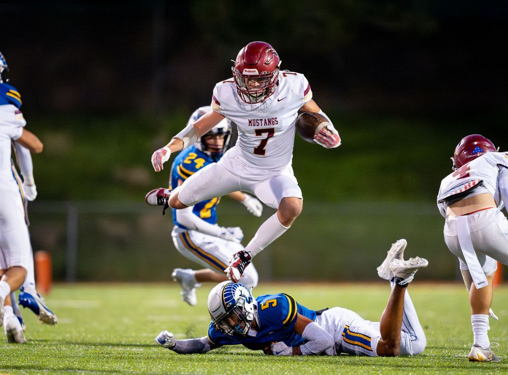 Football player in white jersey leaps over a fallen defender during a night game on the field.