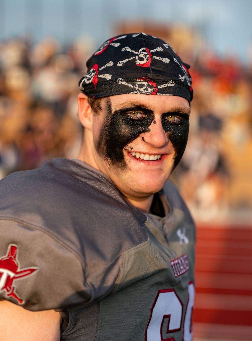 A person wearing a black bandana and sunglasses smiles while wearing a gray jersey at an outdoor sporting event.