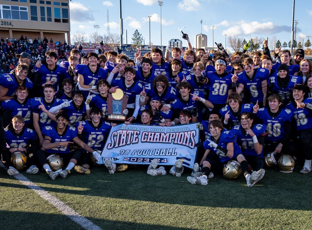 Championship football team poses with trophy banner on field in blue uniforms.