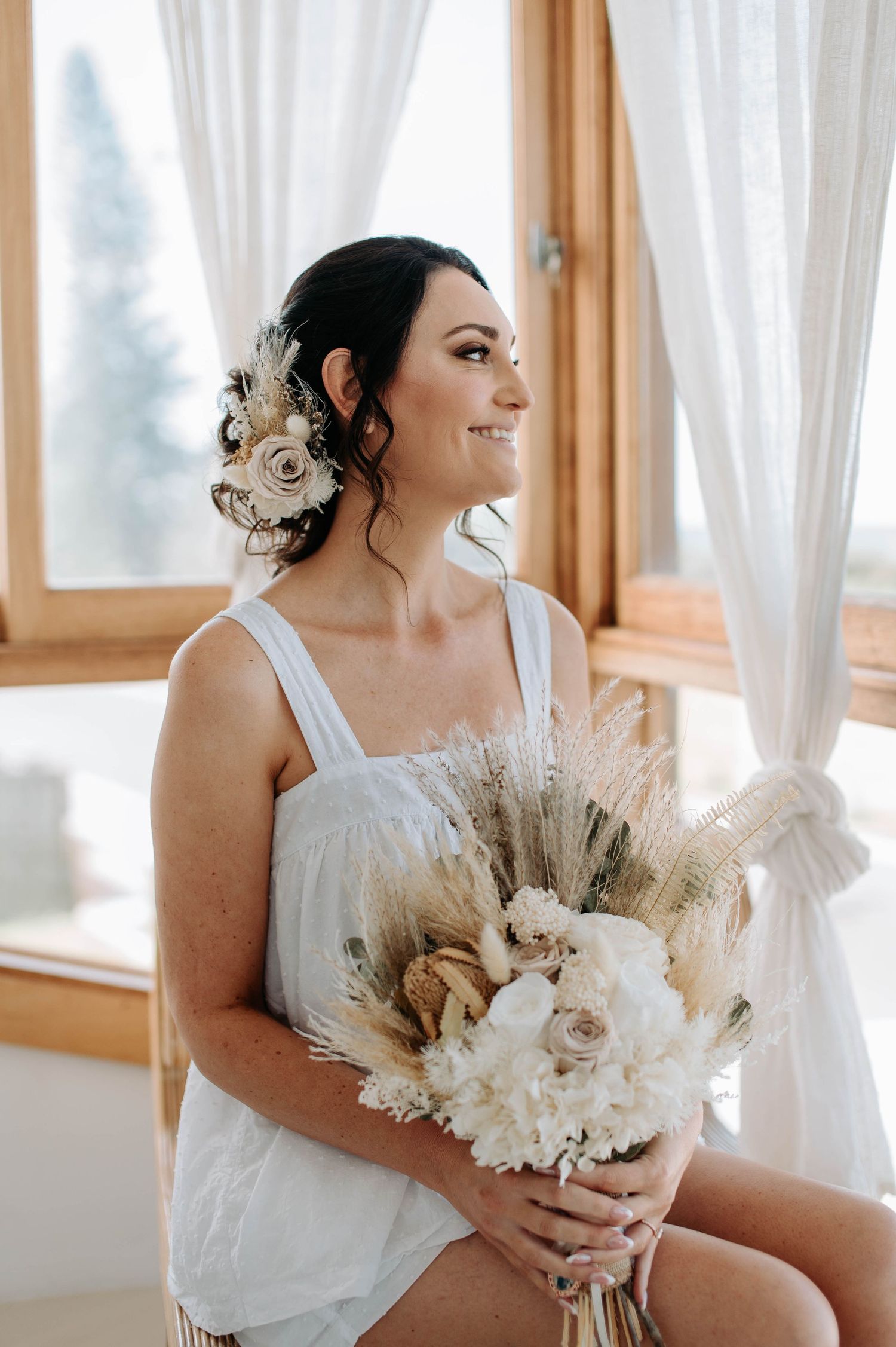 Person in white dress holding dried flowers and pampas grass bouquet by window.